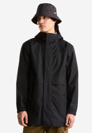 Ung mand iført en sort vandtæt jakke og matchende sort bucket hat, der kigger til siden mod en ensfarvet baggrund.