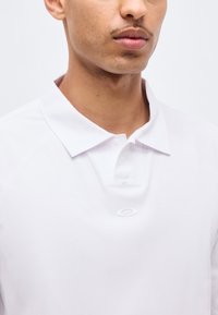 Gros plan sur une personne portant un polo blanc à col avec un logo ovale subtilement embossé sur la poitrine.