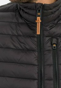 Primer plano de una chaqueta negra acolchada con dos cremalleras verticales, una con un tirador de cuero marrón y la otra con un tirador de tela negra.
