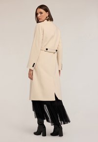 Cappotto di lana beige con un design strutturato, dotato di una vita cintrata, bottoni neri e spacco sul retro. Abbinato a una gonna nera con frange e stivali.