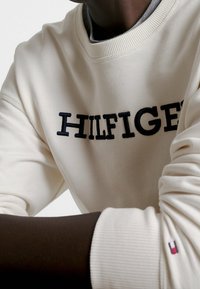 Felpa color crema con ricamo blu navy che recita "HILFIGER". Presenta un colletto e polsini a costine, con un piccolo dettaglio del logo sul manico.