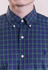 Knapplunghängd skjorta i blå och grön rutigt mönster. Har en klassisk krage, vita knappar och en liten broderad logotyp på vänster sida.
