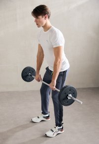 Vægtstang med vægtplader holdt af en person i en tætsiddende hvid skjorte og grå træningsbukser, som udfører et dødløft i et minimalistisk rum.