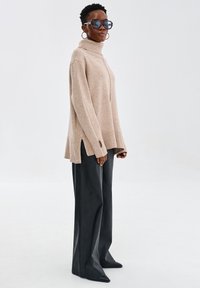 Beżowy, oversize sweter z golfem i bocznymi rozcięciami, zestawiony z czarnymi spodniami typu wide-leg oraz spiczastymi czarnymi butami. Dodatki to duże okulary przeciwsłoneczne.