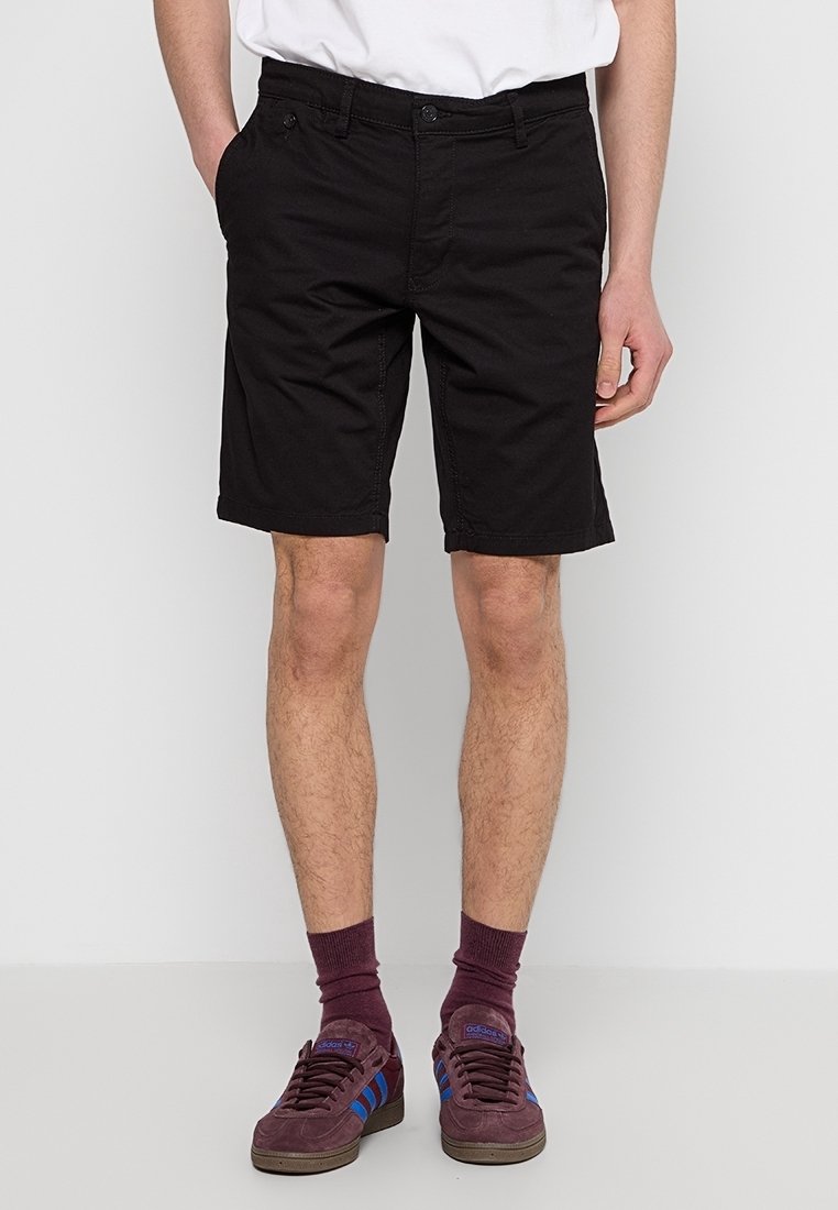 Homme portant un short noir aux genoux, une chemise blanche, des chaussettes bordeaux et des baskets bordeaux avec des bandes bleues, debout devant un fond blanc.