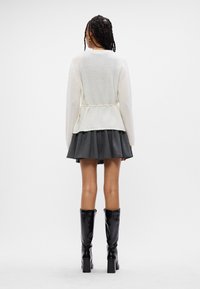 Bílý pletený svetr s peplum designem, v kombinaci s tmavou plisovanou sukní a černými kozačkami na vysokém podpatku. Jednoduché, texturované materiály.