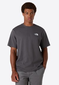 Ung mand står med hænderne i lommen, iført en mørkegrå t-shirt fra The North Face og matchende bukser, vender fremad med neutral ansigtsudtryk.