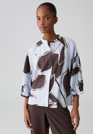 Vrouw draagt een witte blouse met grote abstracte donkergroene bladprints en donkergroene broek, staand tegen een lichtgrijze achtergrond.