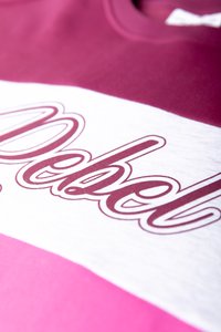 Bordový mikina s bielymi a ružovými horizontálnymi pruhmi, s výrazným bordovým kurzívou textom "Rebel", zobrazená zblízka.