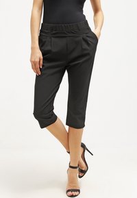 Pantalones recortados negros con un frente plisado y cinturilla elástica, presentando un diseño ajustado y puños enrollados. Combinados con tacones altos negros.