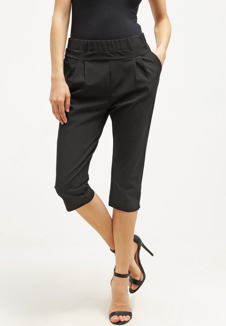 Pantalones recortados negros con un frente plisado y cinturilla elástica, presentando un diseño ajustado y puños enrollados. Combinados con tacones altos negros.