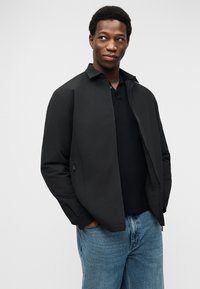 Άνδρας με τα χέρια στις τσέπες, φοράει μαύρο μπουφάν, μαύρο πουκάμισο και μπλε τζιν, κοιτάζοντας πλάγια μπροστά σε απλό φωτεινό φόντο.