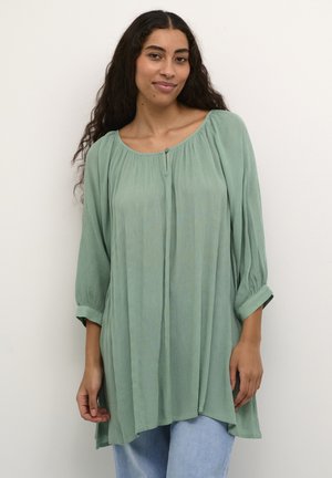 Blouse vert clair texturé avec un col rond et des manches trois-quarts, dotée d'un bouton central et d'une coupe décontractée.