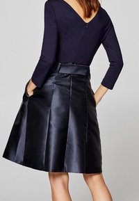 Kvinna i en svart långärmad V-back topp instoppad i en blank svart A-linjekjol med sidofickor, stående framför en enkel grå bakgrund.