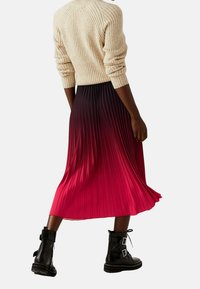 Faltenrock in Midilänge, der sich von Schwarz zu lebendigem Rot verfärbt, kombiniert mit schwarzen Combat-Stiefeln mit Schnallenakzenten und einem strukturierten cremefarbenen Pullover.