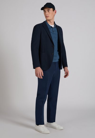 Mężczyzna w granatowej marynarce, niebieskiej koszulce polo, granatowych spodniach, białych tenisówkach i czarnej czapce z daszkiem, stojący na tle szarego tła, patrzący bokiem.