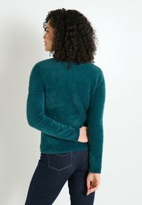 Suéter de lana de color verde azulado, con mangas largas, escote redondo y textura suave, combinado con jeans oscuros. La modelo tiene la espalda hacia la cámara.