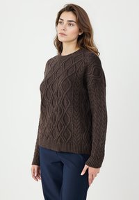 Brązowy sweter dzianinowy z fakturowanym wzorem w warkocze, okrągłym dekoltem oraz ściągaczami u rękawów i na dole. W połączeniu z granatowymi spodniami.