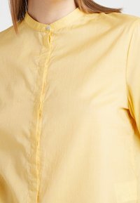 Camicia gialla con colletto, caratterizzata da una chiusura con bottoni, una texture morbida e un tessuto leggero e arioso; nessun motivo o accentuazione visibile.
