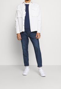 Vit bomullsskjorta med knäppning, två bröstfickor, lager-på-lager över en marinblå sweatshirt. Bärs med blå denimjeans och vita sneakers.