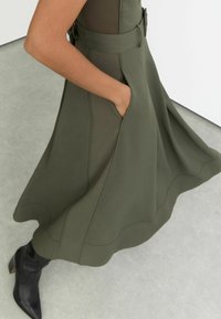 Oliiviroheline kleit struktureeritud disainiga, millel on vöö ja küljetaskud. Sile kangas, lõhvestatud äärega ja peente õmbluse detailidega.