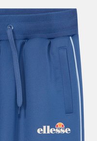 Pantalones deportivos azules con cintura elástica, cordón, bolsillo lateral y raya lateral azul claro. Presenta impresión de logotipo en blanco.