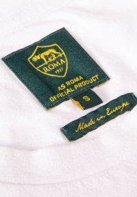 Een witte stoffen label heeft een donkergroene schild met een geel embleem en de tekst "A.S. Roma Officieel Product," maat "S," en "Gemaakt in Europa."