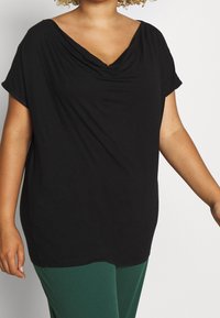 Svart V-ringad topp med draperad halsringning, korta ärmar, avslappnad passform, tillverkad av mjukt tyg, bärs med gröna byxor.