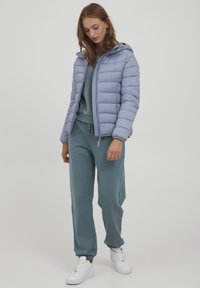 Veste matelassée bleu clair avec une capuche, des poignets côtelés et une fermeture éclair �à l'avant, portée sur un sweat-shirt bleu canard et un pantalon assorti. Des baskets blanches complètent la tenue.