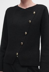 Pull noir à manches longues avec une fermeture asymétrique devant ornée de cinq boutons dorés brillants.