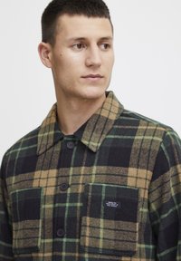 Geruite shirt in zwart, groen en beige flanel. Heeft een knoopsluiting aan de voorkant, grote borstzakken en een spreidkraag. Textuurstof.