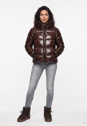 Frau steht mit den Händen in den Taschen, trägt eine glänzende dunkelbraune Steppjacke, hellgraue Jeans und dunkelbraune Schnürstiefel vor weißem Hintergrund.