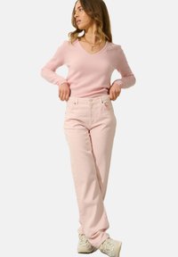 Frau, die ihre hochgeschnittenen hellrosa Jeans anpasst, trägt einen sanften rosa V-Ausschnitt-Pullover und cremefarbene Sneakers, steht vor einem schlichten Hintergrund.