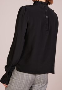 Blusa nera con colletto arricciato e maniche lunghe, caratterizzata da chiusura a bottoni sul retro e polsini aderenti. Tessuto liscio e leggero.