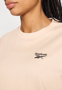 Hanorac Reebok de culoare piersică, cu guler rotund, având un logo negru brodat și o textură de țesătură netedă și moale.