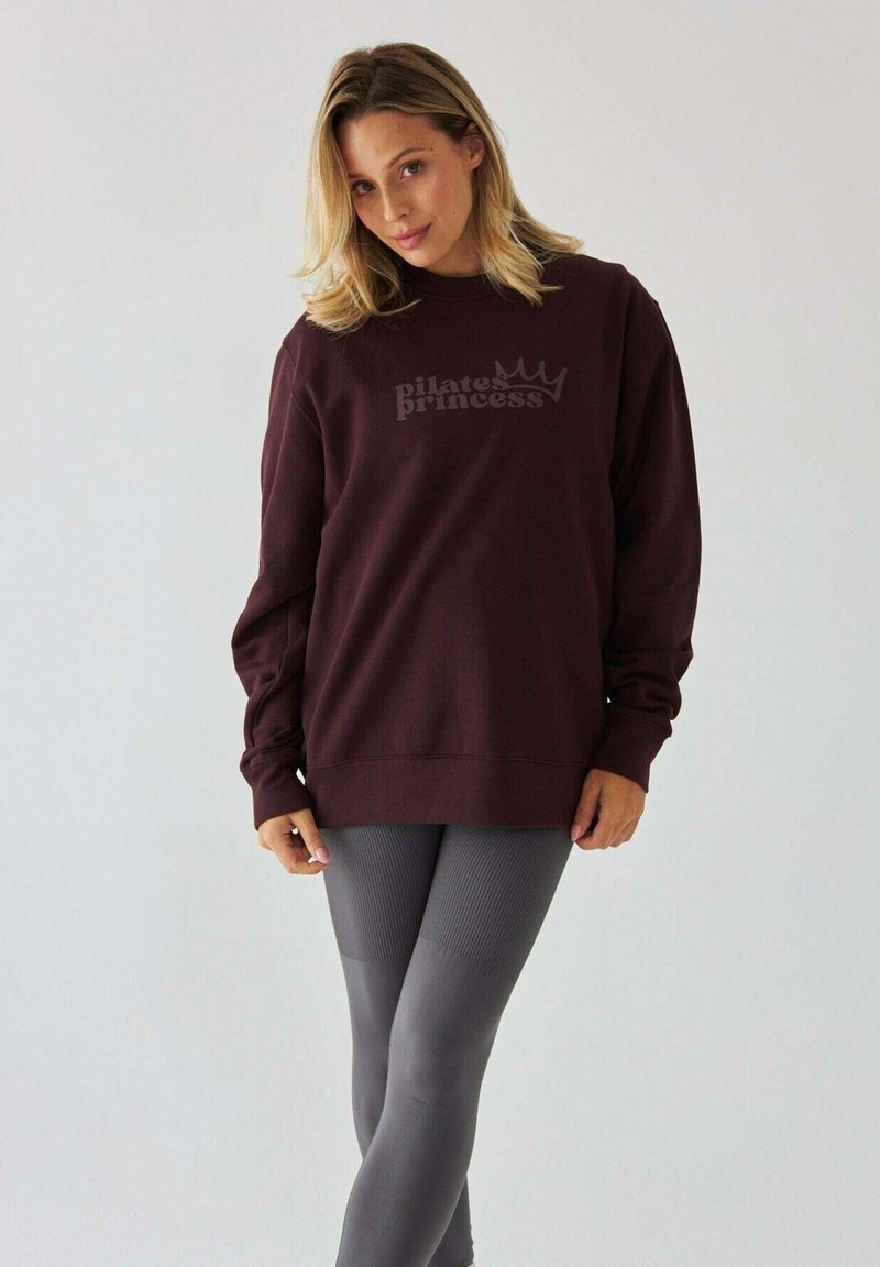 Bordowy sweatshirt z logo "Pilates Princess", długie rękawy, dziane mankiety, w zestawie z szarymi, prążkowanymi legginsami. Gładka faktura materiału.