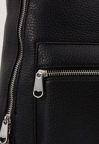 Sac en cuir noir avec surface texturée, fermetures éclair en argent et une poche avant. La tirette de la fermeture éclair comporte un détail découpé en cercle.