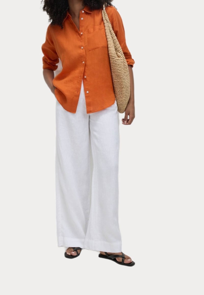 Femme portant une chemise orange à boutons, un pantalon blanc à jambes larges, des sandales noires et portant un grand sac à bandoulière tressé.