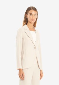 Ljust beige skräddarsydd blazer med en enkel knappstängning, spetsiga slag och sidofickor; slätt tyg och strukturerad design.