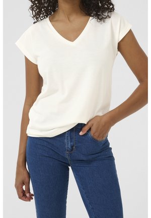Femme portant un t-shirt blanc à manches courtes et col en V, légèrement rentré dans un jean bleu taille haute, avec une main dans la poche et des cheveux bouclés.