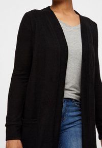 Svart cardigan i mjukt material, med långa ärmar, sjalkrage, öppen front och två sidfickor, överlagd på en grå topp.