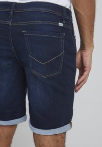Shorts en denim foncé avec ourlets retroussés, dotés d'une poche arrière avec un motif en chevron et des coutures jaunes. Aucun logo de marque visible sur la ceinture.