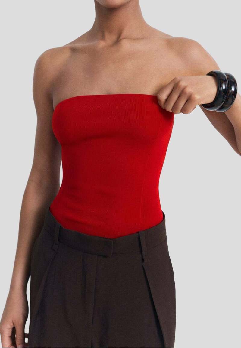 Röd ribbad bandeau-top med en figurnära design, matchad med högmidjade mörkbruna byxor. Modellen justerar toppen.