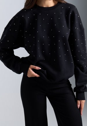 Femme portant un haut noir à manches longues avec petits pois blancs associé à un pantalon noir, main posée sur la hanche sur fond uni.