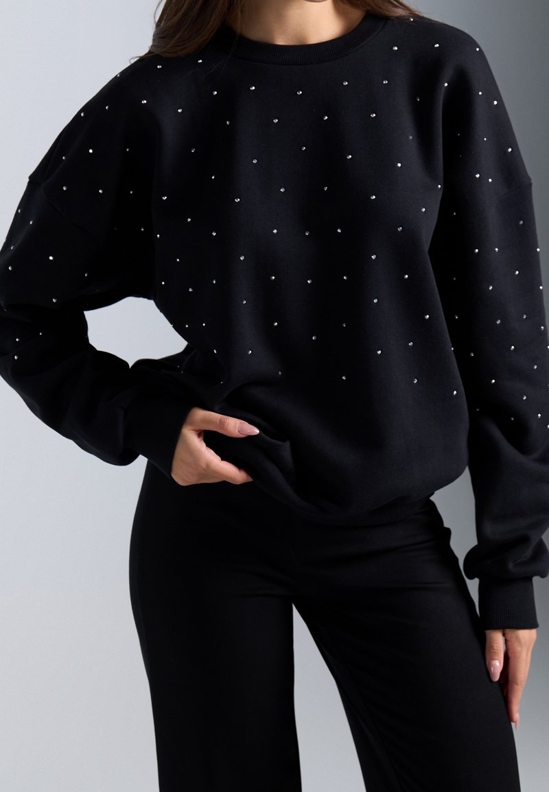 Femme portant un haut noir à manches longues avec petits pois blancs associé à un pantalon noir, main posée sur la hanche sur fond uni.