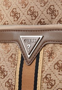 Sac en tissu monogrammé beige avec des accents marron ; présente un logo en argent en forme de triangle, une surface texturée et des détails rayés.