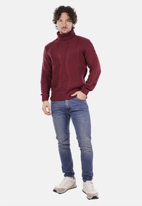 Maglione bordeaux a treccia con collo alto, dalla texture spessa, con polsini a costine, indossato con jeans azzurri e sneakers grigie su uno sfondo neutro.