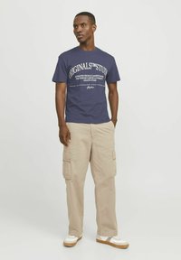 Marineblauw katoenen t-shirt met witte grafische print, gecombineerd met beige cargobroek met zijzakken en een losse pasvorm. De schoenen zijn witte sneakers.