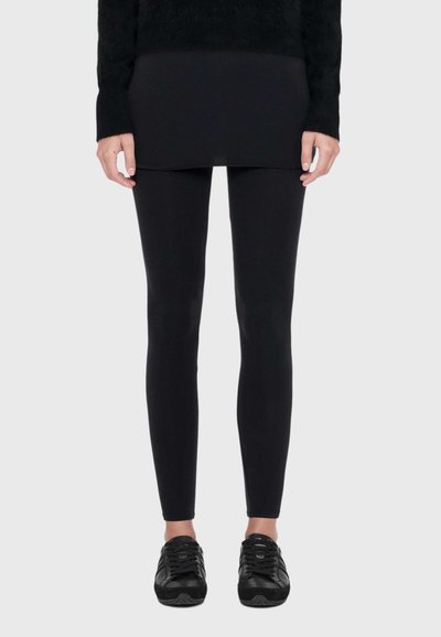 Modèle portant un legging noir, un haut noir long et des baskets noires sur un fond uni clair.