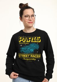 Zwarte sweatshirt met een grafische print van een blauwe auto en gele tekst met de woorden "PARIS STREET RACER." Zacht materiaal met een relaxte pasvorm.